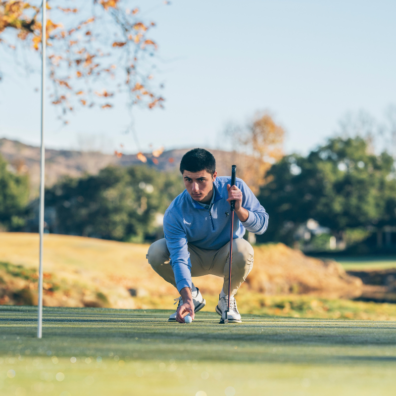 A person crouching on a golf course, aligning a golf ball next to a flagstick while holding a golf club.