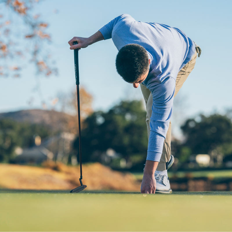 A person in a blue shirt and khaki pants is bending down on a golf course, placing a golf ball on the grass while holding a putter.