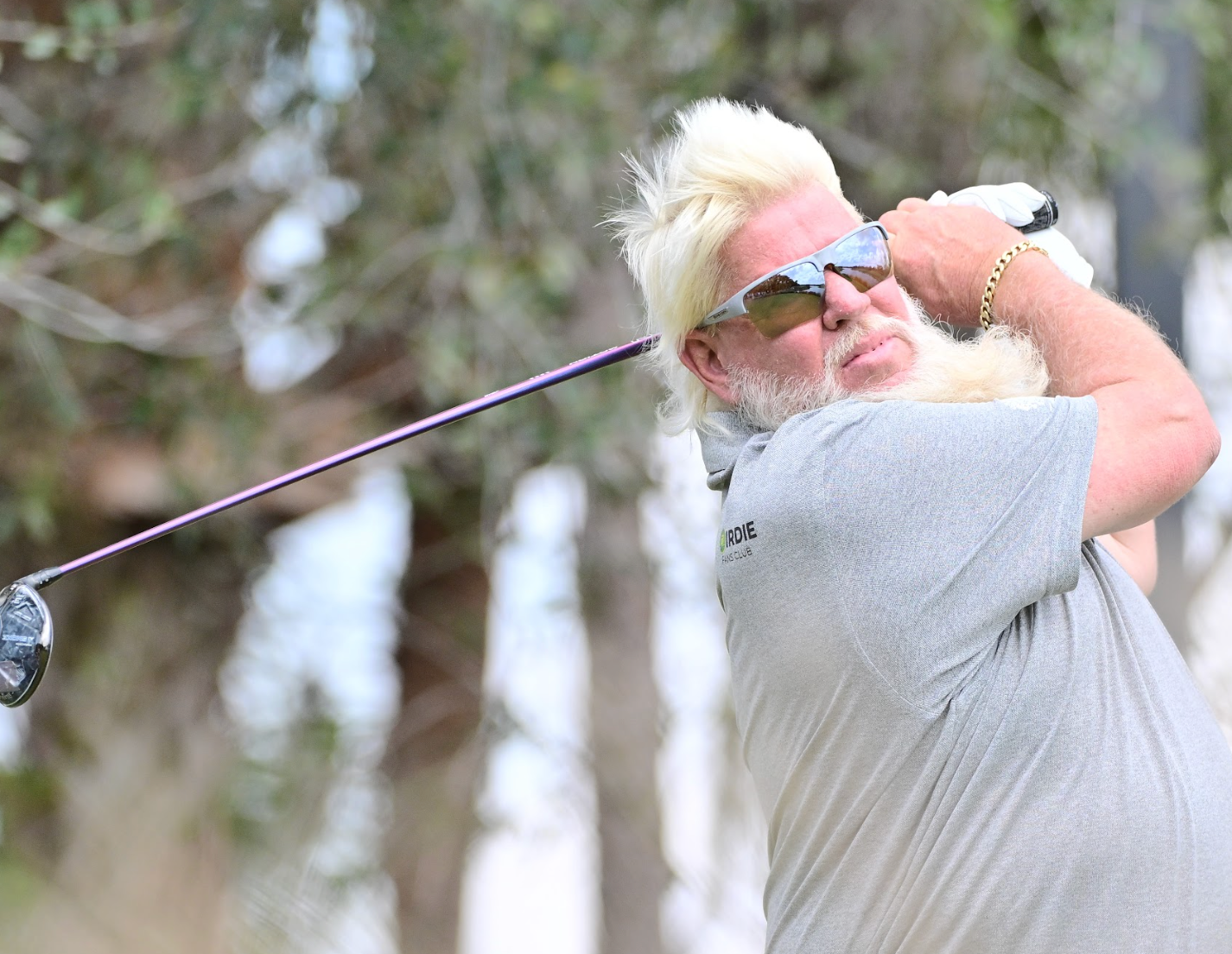 A person with light-colored hair and sunglasses is swinging a golf club outdoors.