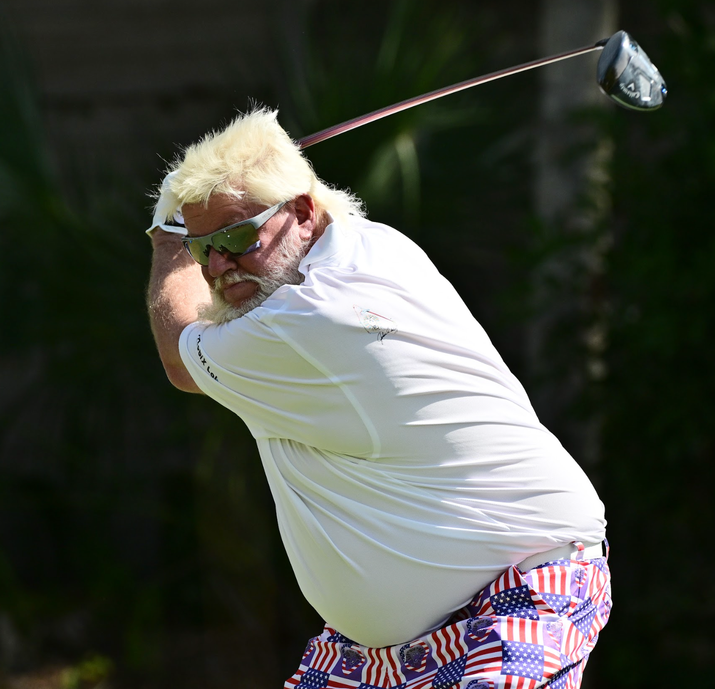 A golfer with sunglasses swings a golf club on a sunny day.