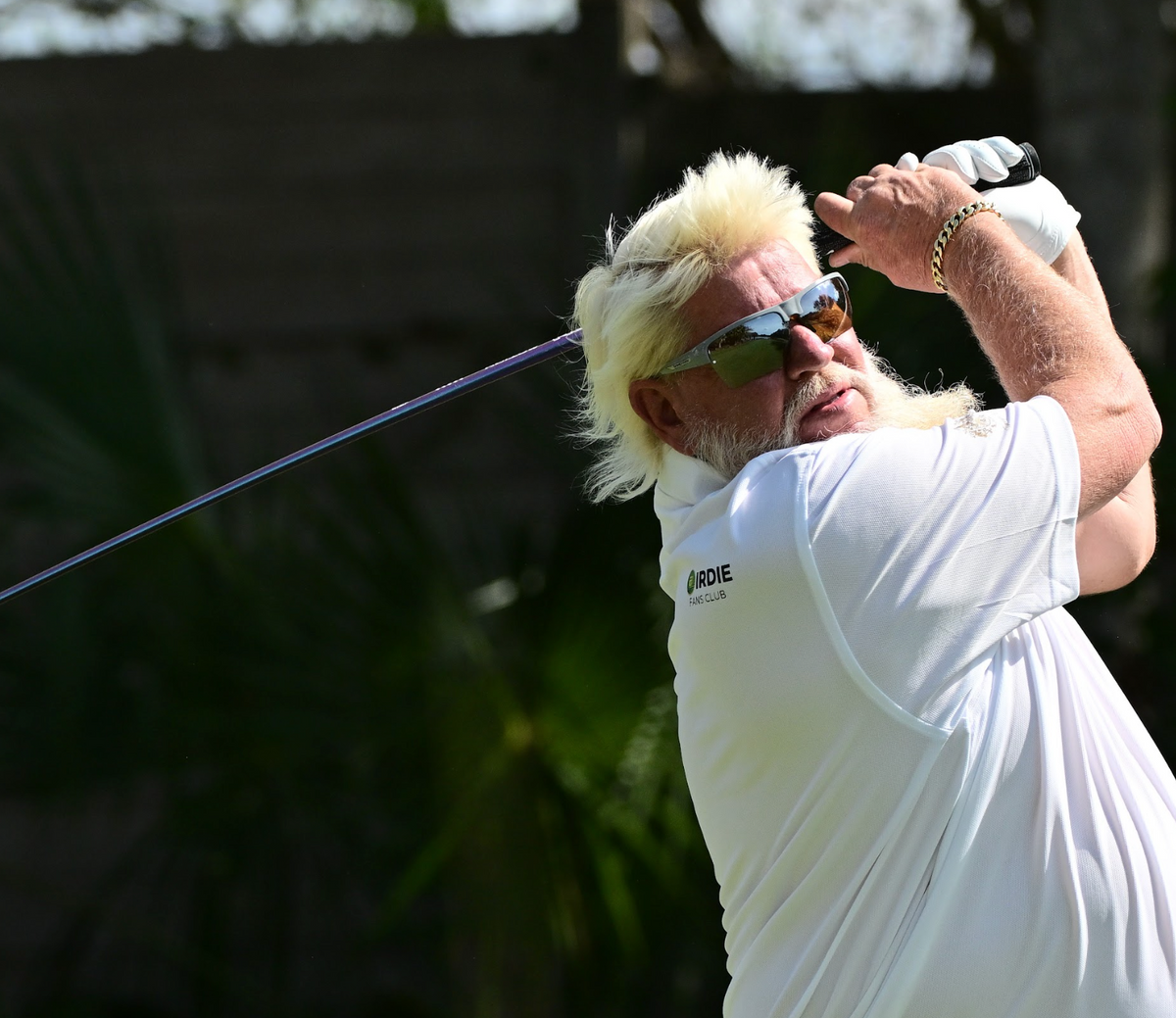 A person with blonde hair and sunglasses is swinging a golf club outdoors.