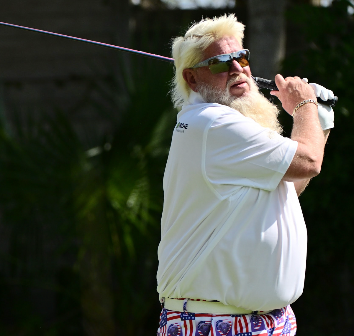 A person with a white beard and sunglasses swings a golf club.