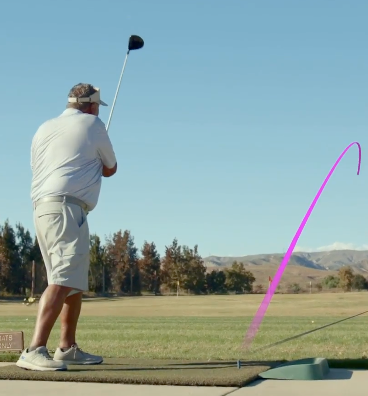 A person is playing golf, swinging a club on a driving range with a purple trajectory line showing the ball's path.
